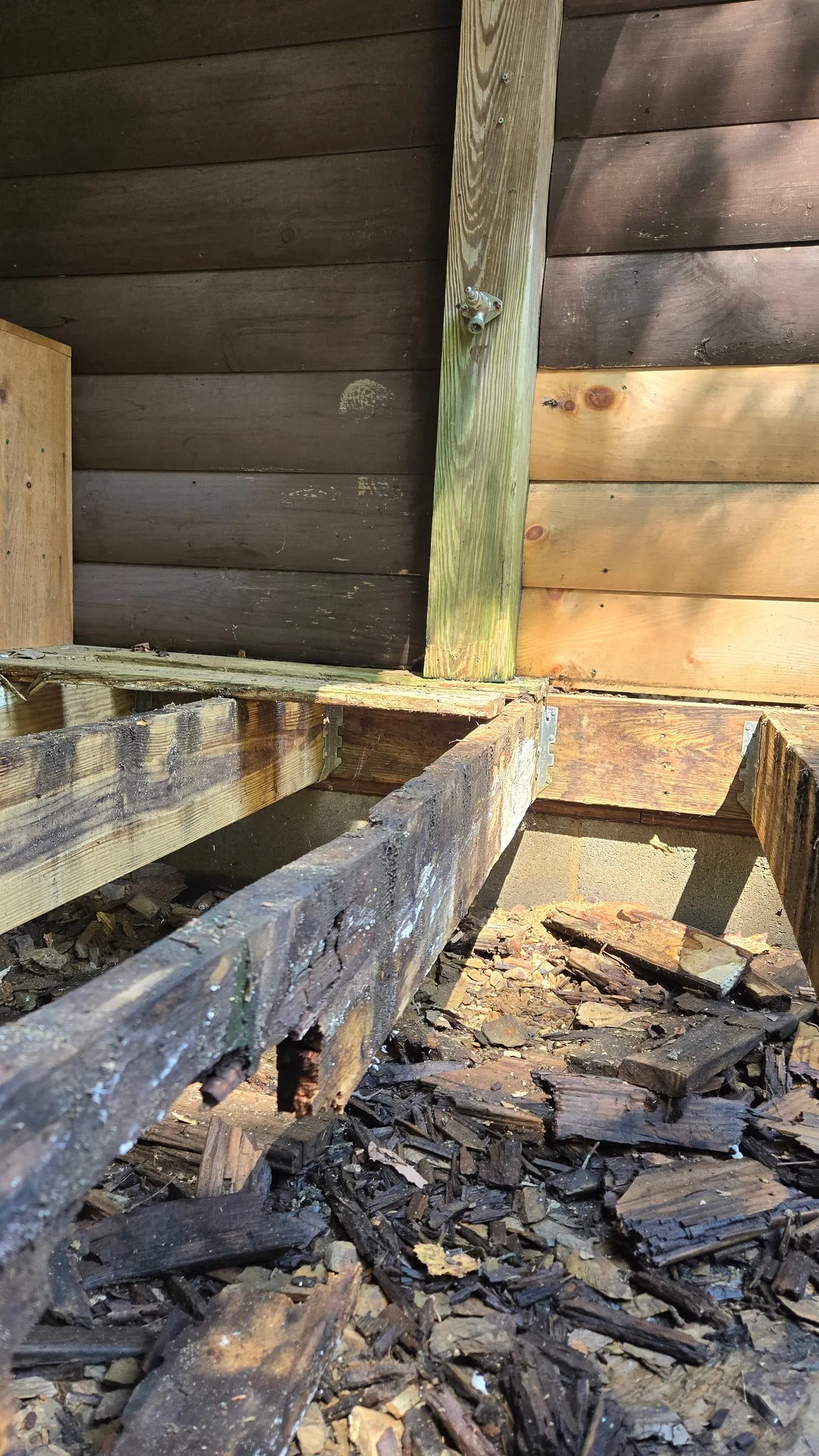 Deck joists in metal hangers at the cabin wall, one with a chunk rotted out of it, rotted debris piled on the ground below