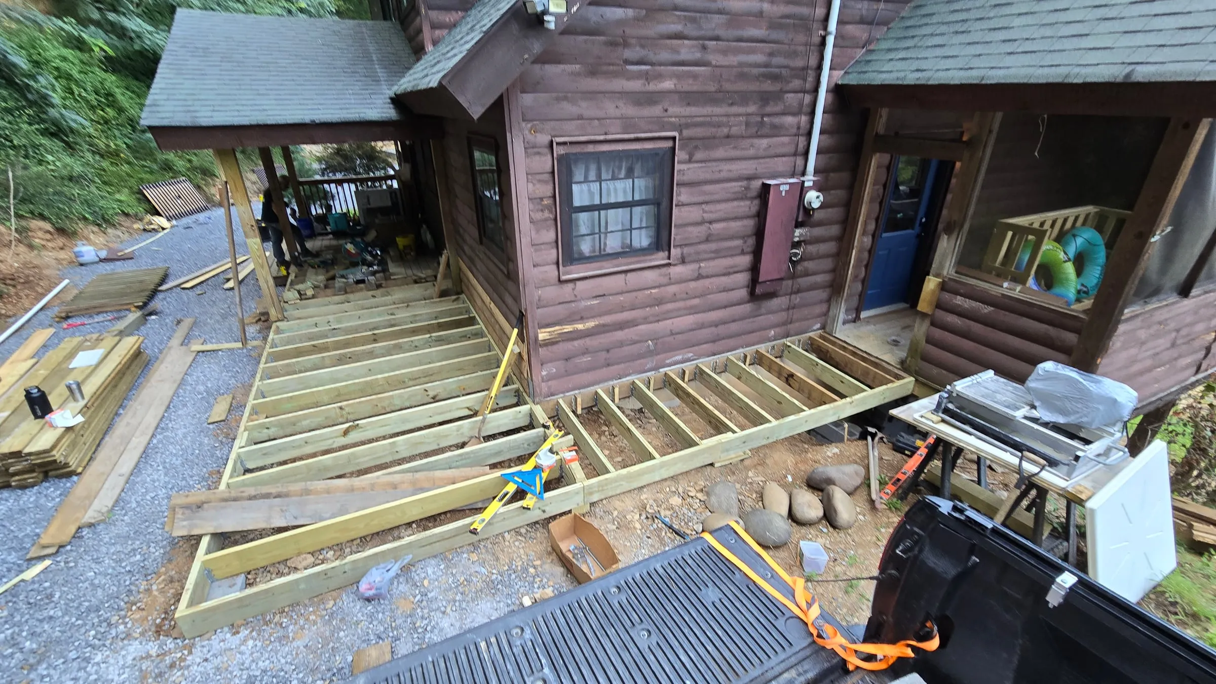 New pressure-treated deck framing with metal joist hangers wrapping two sides of the log cabin, stacks of new decking boards nearby